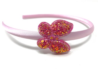 Può includere: Una fascia per capelli rosa chiaro con finitura satinata. Presenta una farfalla scintillante e glitterata nei toni del rosa e dell'oro. Questo accessorio per capelli è perfetto per aggiungere un tocco di fantasia a qualsiasi acconciatura.