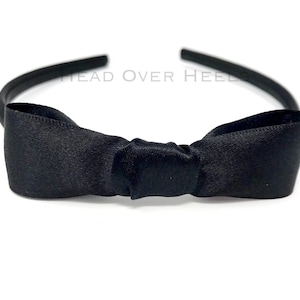 Peut inclure: Un serre-tête noir avec un nœud en satin. Le nœud est noué au centre et orne le devant du serre-tête. Le texte "HEAD OVER HEELS" est visible sur le serre-tête.