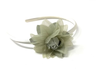 May include: A light-colored headband with a large, layered flower in shades of green and gray. The flower is the focal point, attached to a thin, satin-covered band. The accessory is set against a white background.