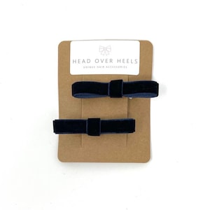 May include: Two navy blue velvet bow hair clips on a brown card with the text "HEAD OVER HEELS UNIQUE HAIR ACCESSORIES". The bows are rectangular with a small square knot in the center.
