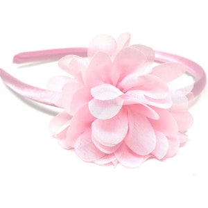 Pode incluir: Uma tiara rosa claro com uma flor grande e em camadas. A flor é feita de pétalas de tecido transparente e delicado. A tiara em si é uma faixa sólida coberta de cetim. Acessório de cabelo.