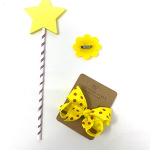 May include: Yellow star-shaped wooden wand with purple and white stripes, a yellow felt flower with a pin, and a yellow bow with brown polka dots on a brown card with the text "Head Over Heels Unique Hair Accessories".