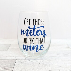 May include: A clear glass wine glass with a blue glitter design that says "Get those meters drink that wine".
