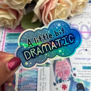 Op de afbeelding: Een holografische sticker in de vorm van een blauwe wolk met de tekst "A little bit DRAMATIC" in zwarte letters en witte sterren.