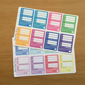 May include: A set of 12 colorful stickers with a checklist for healthy options. Each sticker has a different color and includes the letters BLD, a space for writing, and a section for checking off healthy options. The stickers are designed for use in a planner or journal.