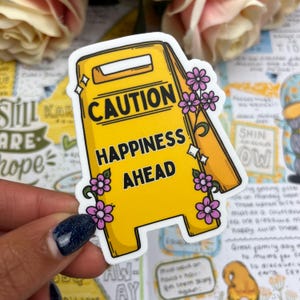 May include: Yellow caution sign sticker with the text "Caution Happiness Ahead" and pink flowers around the sign.
