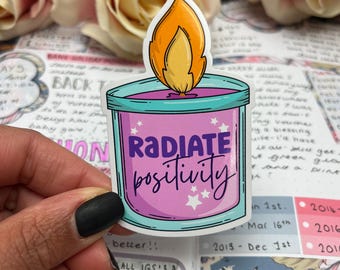 May include: A sticker featuring a cartoon candle with a flame. The candle is in a teal container with a pink interior and the words "RADIATE positivity" in purple. White stars are scattered around the text. The background includes a journal and roses.