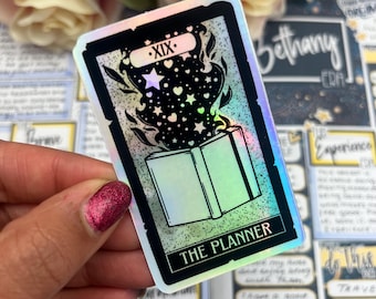 May include: A holographic sticker featuring a tarot card design. The card has a black border and depicts an open book with stars and hearts emanating from it. The text "XIX" and "THE PLANNER" are visible.