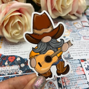 May include: A sticker depicting a cartoon gnome in a brown cowboy hat and boots, playing a guitar. The gnome has a grey beard, yellow shirt and blue jeans. The sticker is set against a floral background.