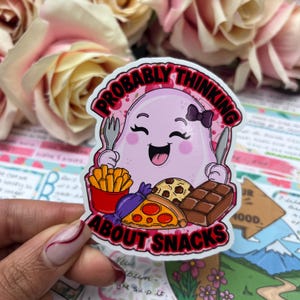 Pode incluir: Um autocolante com um personagem fantasma rosa segurando um garfo e uma faca, rodeado de lanches. O texto "PROBABLY THINKING ABOUT SNACKS" está em vermelho e branco. Os lanches incluem batatas fritas, pizza, chocolate e um biscoito.