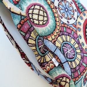Könnte beinhalten: Eine bunte Tapisserie-Tasche mit einem geometrischen Muster aus Kreisen und Blumen. Die Tasche hat einen langen Riemen und ein kleines Etikett mit der Aufschrift "orig."
