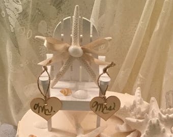 May include: A three-tiered wedding cake decorated with white frosting, seashells, and starfish. The top tier features a miniature white wooden beach chair with a starfish and a bow. The chair has two signs hanging from it that say "Mr." and "Mrs."