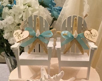 May include: A two-tiered wedding cake decorated with white frosting and a blue ribbon. The top tier features two miniature white Adirondack chairs with starfish and blue bows. The chairs have signs that say "Mr." and "Mrs."