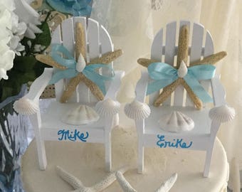 May include: A two-tiered white wedding cake decorated with white starfish and seashells. The top tier has two white miniature beach chairs with blue bows and the names "Mike" and "Erika" written on the front.