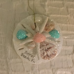 May include: White sand dollar ornament with a pink and white seashell, blue and white seashells, and gold glitter. The ornament has the text "Delray Beach, Fl" written on it.