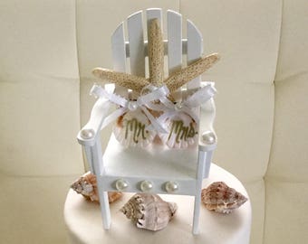 May include: A two-tiered white wedding cake decorated with seashells and starfish. The top tier features a miniature white wooden chair with a starfish couple sitting on it. The chair is decorated with pearls and the words "Mr." and "Mrs."