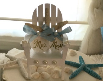 May include: A white wedding cake with a white wooden beach chair topper. The chair has a blue ribbon and two seashells that say "Mr. Mrs." The cake is decorated with white seashells and two starfish, one blue and one white.