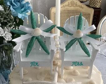 May include: A white wedding cake topped with two miniature Adirondack chairs, each adorned with a teal starfish and a seashell. The chairs are labeled "Mr." and "Mrs." in teal script. Seashells and a starfish decorate the cake's surface.