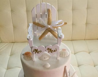 May include: A three-tiered wedding cake decorated with seashells, starfish, and a white wooden chair with a starfish on top. The cake is decorated with a silver ribbon and the words "Mr." and "Mrs." are on the top tier.