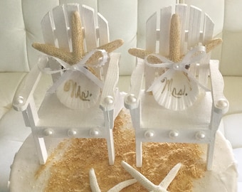 May include: A wedding cake decorated with a beach theme. Two miniature white Adirondack chairs are adorned with starfish and seashell name tags that read "Mrs.". Additional starfish and sand-like sprinkles complete the design.