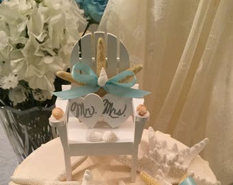 May include: A white wedding cake decorated with seashells and starfish. The top of the cake has a white wooden chair with a starfish and a blue bow. The chair has two signs that say "Mr." and "Mrs."