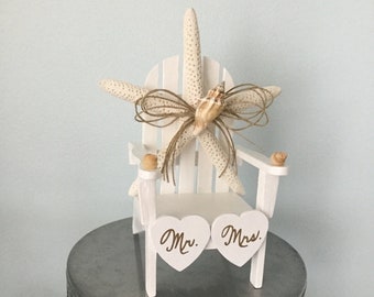 May include: White wooden chair with a starfish and seashells on top. The chair is on a silver cake stand with a glass base. The chair has two white hearts attached to the front, one says "Mr." and the other says "Mrs."
