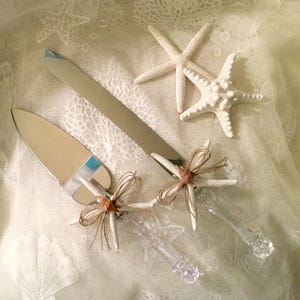 May include: A set of two silver cake servers with clear plastic handles decorated with starfish and twine bows. The servers are on a white lace tablecloth.