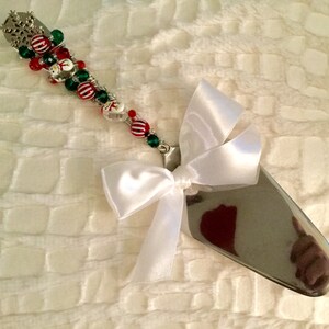 May include: A silver cake server with a white satin bow and a beaded Christmas-themed handle. The beads are red, green, and white and include snowman charms.