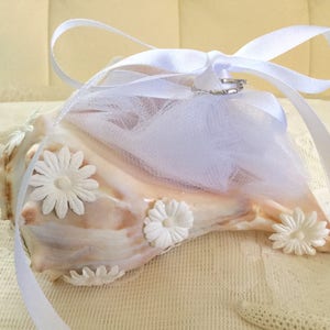 May include: A seashell ring bearer pillow with white fabric flowers and a white tulle overlay. The pillow is tied with a white satin ribbon.