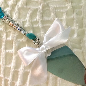 May include: A silver cake server with a teal blade and a handle adorned with blue beads and colorful letter beads spelling out a name. A white satin bow is tied near the blade.