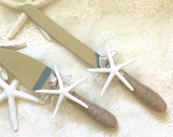 May include: A pair of white cake cutting knives with burlap handles decorated with starfish and seashells. The knives are on a white lace tablecloth.