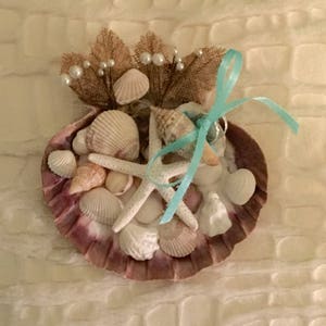 May include: A seashell ring holder with a starfish, seashells, and a turquoise ribbon. The ring holder is decorated with burlap and pearls.