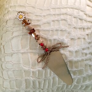 May include: A silver cake server with a beaded handle. The handle is decorated with a white daisy, brown, red, and gold beads.
