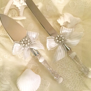 May include: A wedding cake knife and server set. The stainless steel blades are paired with clear acrylic handles. Each handle is adorned with a white bow, pearl accents, and a cluster of rhinestones. The set is displayed on a cream-colored surface with seashells.