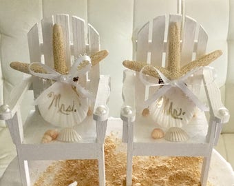 May include: White wooden chairs with starfish and seashells, decorated with white ribbon and the words "Mr." and "Mrs." on the shells. The chairs are on a sandy surface.