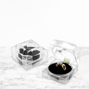 May include: Two clear plastic ring boxes. One box is open and shows a gold ring with a single clear stone in the center. The ring is sitting on a black velvet cushion.