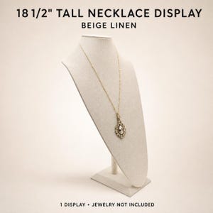 May include: An 18 1/2" tall necklace display stand covered in beige linen fabric. The display features a slanted neck and chest form on a rectangular base. Text at the top reads 18 1/2" TALL NECKLACE DISPLAY BEIGE LINEN and at the bottom 1 DISPLAY.