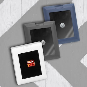 May include: Three small, square, black velvet display boxes with clear lids. Each box contains a different gemstone: a clear diamond, a clear diamond, and a red gemstone.