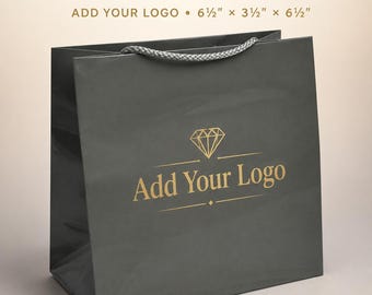 May include: A glossy grey paper bag with a braided grey cord handle. The bag features a gold diamond icon and the text Add Your Logo. The text 100 CUSTOM GLOSSY GREY BAGS ADD YOUR LOGO 6 1/2 inches x 3 1/2 inches x 6 1/2 inches is printed above.