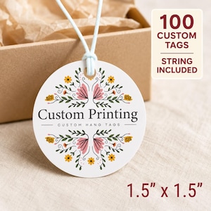 Op de afbeelding: Een rond wit label van 3,8 cm bij 3,8 cm met een lichtblauw touwtje. Het label heeft een bloemmotief in roze, geel en groen, met de tekst Custom Printing en Custom Hang Tags. Een label geeft aan dat het om 100 aangepaste labels gaat.