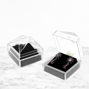May include: Clear acrylic jewelry boxes with black velvet inserts. One box is open, displaying a pair of silver-toned earrings with pink gemstones. The other box is closed. The boxes have a geometric design.