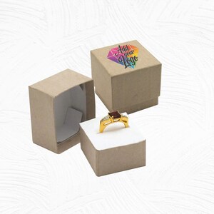 May include: A brown cardboard ring box with a white interior. The box is open and a gold ring with a red stone and clear stones is inside. The box has a lid with a rainbow diamond design and the text "Add your Logo".