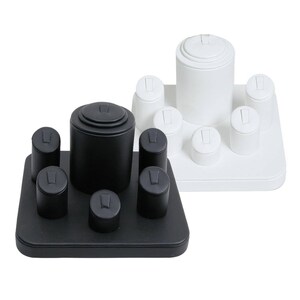 May include: A black and white ring display stand with eight ring holders. The stand is made of a smooth, textured material and has a square base. The ring holders are cylindrical and have a small indentation on top for holding rings.