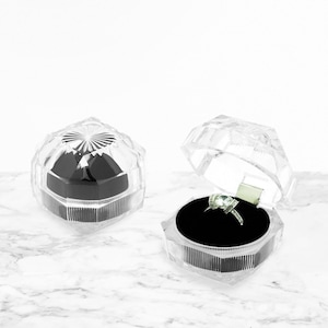May include: Two clear plastic ring boxes with a black velvet interior. One box is open and shows a silver ring with a clear stone.