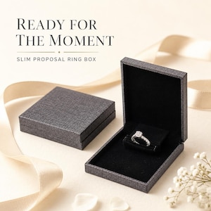 May include: Two slim, dark gray, textured ring boxes with black velvet interiors. One box is closed, and the other is open, displaying a silver-toned ring with a central stone. Text reads: READY FOR THE MOMENT SLIM PROPOSAL RING BOX.