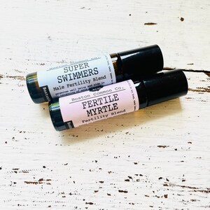 May include: Two small brown glass roller bottles with black caps. The bottle on top has a white label with the text "SUPER SWIMMERS Male Fertility Blend" and the bottle on the bottom has a pink label with the text "FERTILE MYRTLE Fertility Blend".