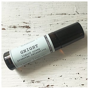 Könnte beinhalten: Eine schwarze und klare Glas-Rollflasche mit einem weißen Etikett, auf dem "GNIGHT liquid sleep promotes relaxation a special blend of essential oils shake well" steht.
