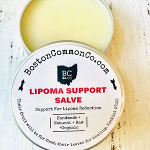 May include: A white tin with a black and white label that reads "BostonCommonCo.com" and "LIPOMA SUPPORT SALVE". The label also includes the text "Support For Lipoma Reduction", "Handmade + Natural + Raw +Organic", and "Their fruit will be for food, their leaves for healing. Ezekiel 47:12".