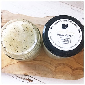 May include: A glass jar of lemon verbena sugar scrub with a black lid. The lid has a white label with the text "Boston CommonCo.com", "Lemon Verbena Sugar Scrub", "handmade + natural", and "Their fruit will be for food, their leaves for healing. Ezekiel 47:12".