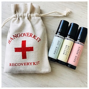 Könnte beinhalten: Eine weiße Stofftasche mit dem Text "Hangover Kit" und einem roten Kreuz. Die Tasche enthält drei kleine Roll-on-Flaschen mit den Etiketten "Knock Out Headache Blend", "Motion Potion Vertigo and Dizziness" und "Have Mercy Sore No More".